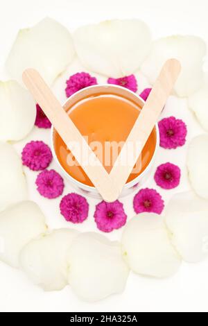 Pot de cire avec bâtonnets de bois, fleurs roses et pétales blancs sur fond blanc.Concept d'épilation.Vue de dessus de la pose à plat Banque D'Images