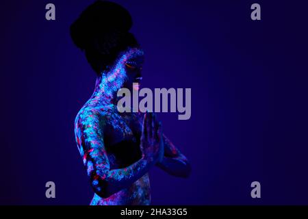 Jeune femme de prière noire mince avec des impressions fluorescentes colorées sur la peau, belle femme priez, demandez la bénédiction. Impressions lueurs dans les rayons UV. Art du corps, Banque D'Images