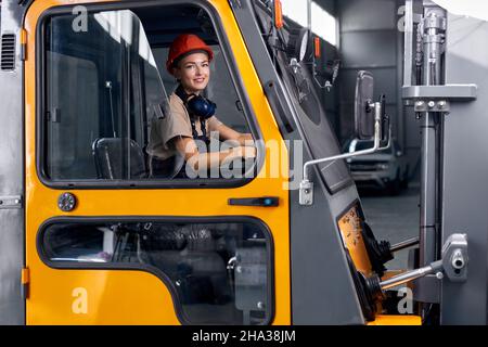 Une employée qui conduit une chargeuse dans un entrepôt industriel, a l'air heureuse, vêtue d'un uniforme d'ingénieur et d'un casque orange rigide, souriant au camiseur Banque D'Images