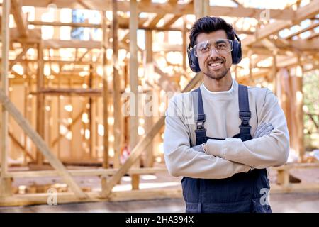 homme caucasien souriant en casque et uniforme au travail, maison en bois.Un jeune homme barbu en lunettes de protection regarde la caméra en tenant les bras repliés Banque D'Images