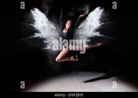 Belle ballerine femme en bodysuit noir danse magnifiquement ballet en studio sombre avec de la farine sur fond noir.Athlète femelle saut avec élégance Banque D'Images