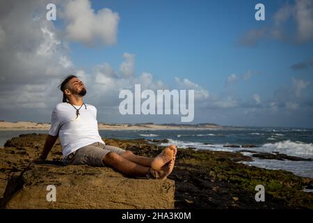 Un jeune homme de race blanche et hippie, adulte, se détend sur les rochers du bord de mer au lever du soleil.Tourisme profitant de la nature. Banque D'Images