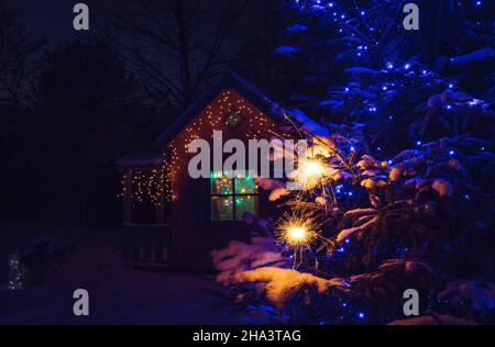 Accent sélectif sur les bougies de sparkler accrochées et brûlantes sur l'épicéa à l'extérieur pendant la nuit hivernale enneigée.Jolie maison de jeu décorée sur fond. Banque D'Images