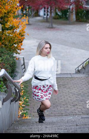 Jeune femme blanche européenne aux cheveux blonds en chandail blanc en jupe fleurie dans une ville grimpant les escaliers Banque D'Images
