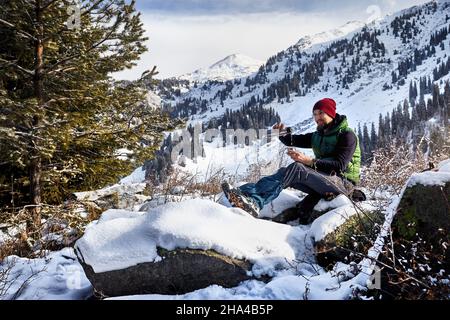 Homme avec une barbe en chapeau rouge qui verse le thé chaud des thermos dans la forêt d'épicéa sur fond de montagne d'hiver. Banque D'Images