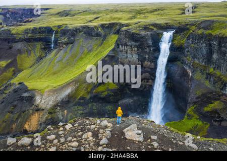 homme en manteau jaune appréciant la cascade de haifoss dans le canyon de landmannalaugar, islande. vue aérienne panoramique de drone. Banque D'Images