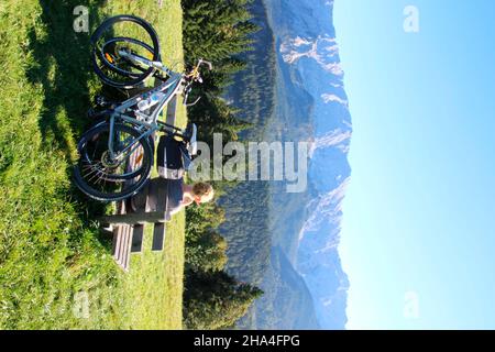 une jeune femme fait une pause lors d'une visite à vélo sur un banc à l'eckbauer en face du massif de wetterstein avec un ciel bleu merveilleux, l'automne dans le pays de werdenfelser, l'europe, l'allemagne, la bavière, la haute-bavière, garmisch partenkirchen, le temps de rêve Banque D'Images