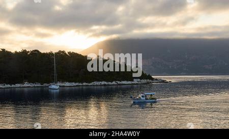 grèce,iles grecques,iles ioniennes,kefalonia,fiskardo,humeur du matin,ciel partiellement nuageux,vue du lever de soleil sur le bassin du port,couverture nuageuse grise,ithaca en arrière-plan,petit bateau de pêche vient d'entrer dans le port,bateau à voile est ancré en face de la pointe Banque D'Images