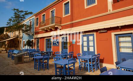 grèce,iles grecques,iles ioniennes,kefalonia,fiskardo,ambiance matinale,ciel partiellement nuageux,café,bar ou restaurant,façade rouge avec lumière matinale devant des tables et chaises bleues vides Banque D'Images