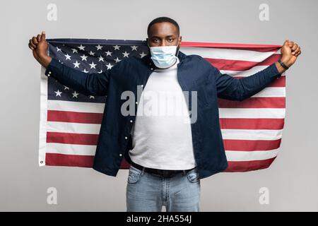 Un jeune homme afro-américain qui milte pour les droits dans un masque médical protecteur tient derrière lui un large drapeau américain ouvert, un homme souriant et joyeux protestant contre le racisme, isolé sur un mur gris Banque D'Images