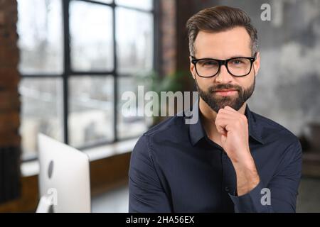 Homme d'affaires masculin réussi en costume formel coûteux, tenant le bras près du menton, a un chaume soigné, confiant, homme riche posant pour la photo, regardant l'appareil photo Banque D'Images