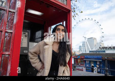 Fille souriante dans le centre de Londres dans un coffret téléphonique anglais rouge typique avec le London Eye derrière elle Banque D'Images