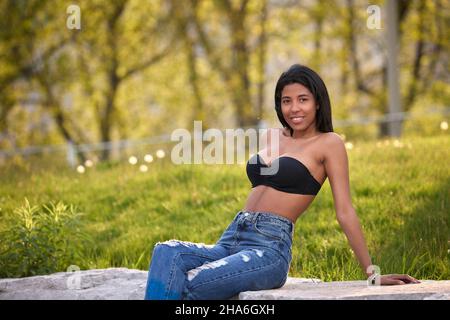 Femme colombienne souriante portant un Jean denim et un haut noir assis à l'extérieur sous la lumière chaude du soleil à Toronto, Canada. Banque D'Images