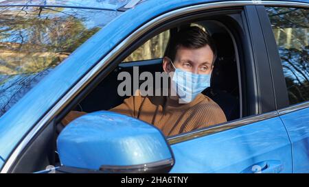 Un homme aux cheveux foncés dans un masque médical est assis en voiture sur le bord de la route près de la forêt Banque D'Images