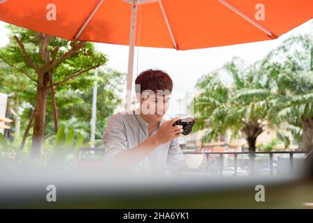 Un jeune homme souriant assis à table dans un café avec un appareil photo tout en buvant une tasse de café Banque D'Images