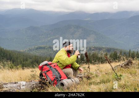 Un mâle caucasien barbu en vacances tout en randonnée dans les montagnes se trouve sur le tronc d'un arbre tombé et de boire du thé ou du café d'un thermos Banque D'Images