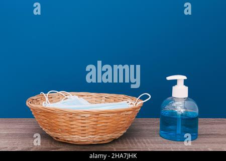 Sur une table en bois devant un fond bleu foncé se trouve un panier en osier avec des masques chirurgicaux et à côté d'un distributeur de savon hydroalcoolique Banque D'Images