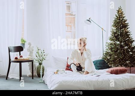 Jeune belle femme dans un intérieur moderne du nouvel an, dans une chambre blanche, sur le fond des lumières de bokeh d'un arbre de Noël.Sélection douce Banque D'Images