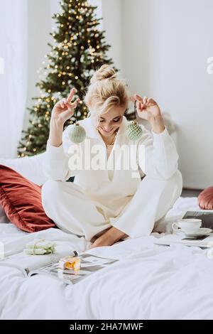 Jeune belle femme dans un intérieur moderne du nouvel an, dans une chambre blanche, sur le fond des lumières de bokeh d'un arbre de Noël avec décoration Banque D'Images
