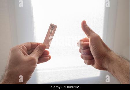 Vue de la première personne des mains d'un homme tenant un test d'antigène Covid-19 dans une main, et de l'autre main avec le pouce vers le haut, faisant le symbole ok, en arrière Banque D'Images