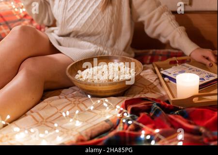 femme sans visage en laine tricotée confortable chandail dans le lit sur une couverture au camping mobile remorque avec un livre et pop-corn nourriture et bougie de détente et en Banque D'Images