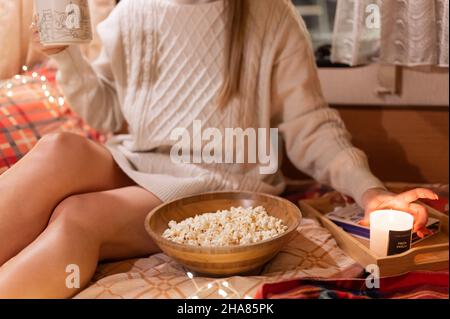 femme sans visage dans un confortable chandail dans le lit sur une couverture au camping remorque mobile à la maison avec une tasse de café ou de thé et pop-corn nourriture et bougie de détente et enj Banque D'Images
