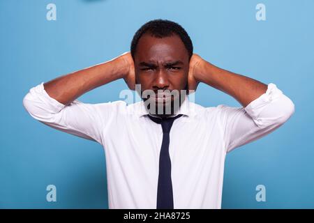 Portrait d'un homme d'affaires en frognant debout couvrant les oreilles pour arrêter d'entendre le bruit voulant le silence.Travailleur de bureau stressé ayant un mal de tête mettant les bras au-dessus de la tête dans un environnement bruyant. Banque D'Images