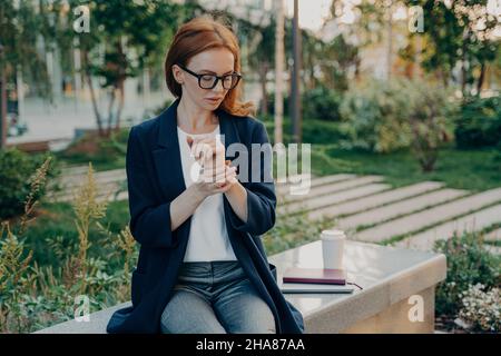 Jeune femme d'affaires à tête rouge fatiguée mesurant le pouls au poignet, vérifiant le rythme cardiaque, assis dans le parc Banque D'Images