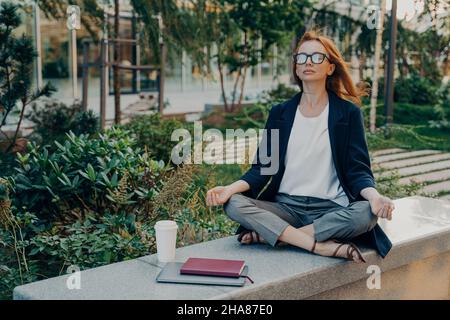 Femme d'affaires paisible tenant les mains dans le geste de mudra, assis dans la pose de lotus et méditant dans le parc Banque D'Images