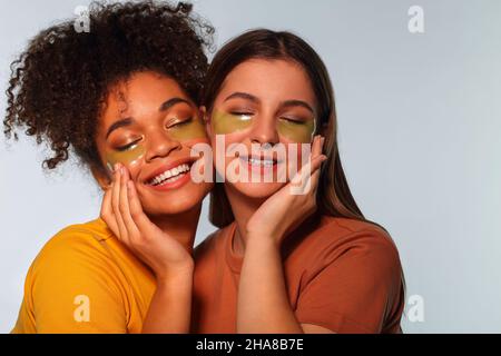 JOURNÉE AU SPA.Deux filles multiraciales avec des taches d'oeil près l'une de l'autre joue à joue, souriant agréablement avec les yeux fermés, amies heureux appréciant sk Banque D'Images