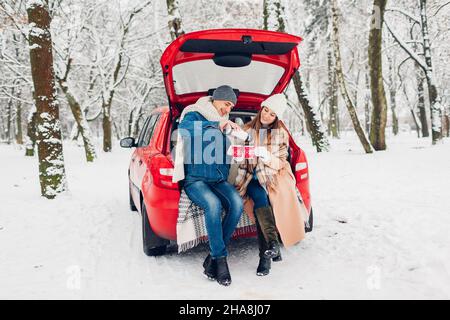 Couple amoureux assis dans le coffre de voiture ayant le thé chaud dans la forêt enneigée d'hiver.Homme qui verse la boisson du thermos.Les gens se détendent à l'extérieur lors d'un voyage en voiture Banque D'Images