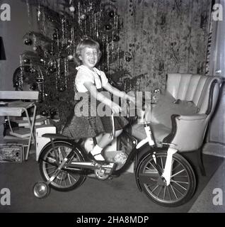 1960s. historique, une jeune fille excitée et ravie assis sur son cadeau de noël, une nouvelle bicyclette. Banque D'Images