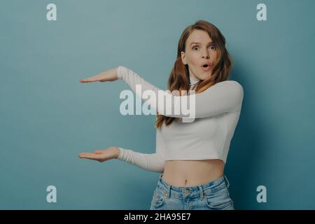 Surprise étonné jeune femme montre quelque chose de grand dans la taille avec les mains tout en se tenant isolé sur bleu Banque D'Images