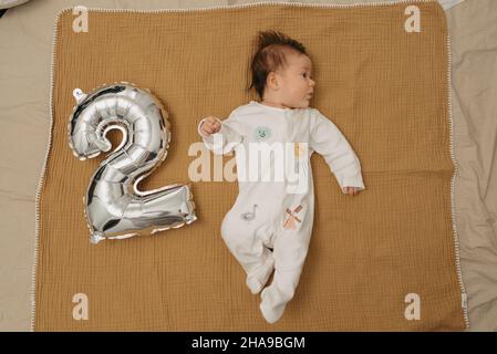 Un bébé se détend sur une couverture en mousseline près d'un ballon en feuille d'argent en forme de 2 et regardant à droite.Une fille de bébé dans un vêtement d'une pièce est Banque D'Images