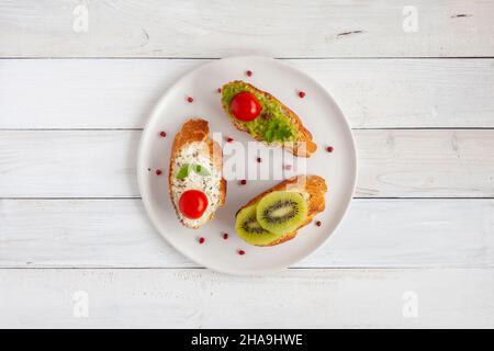 trois mini-sandwiches faits maison avec fromage à la crème et légumes, sur une plaque blanche sur fond de bois, vue du dessus Banque D'Images