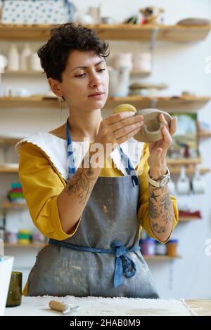 Femme inspirée utilisant une éponge pour absorber l'eau sur des ustensiles de cuisine en argile, visite atelier de céramique Banque D'Images