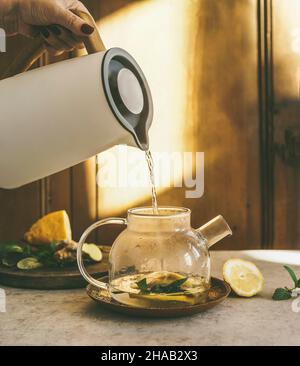 Une femme verse de l'eau chaude à la main dans une théière en verre avec de la menthe, du gingembre et du citron à l'arrière-plan de la cuisine avec lumière naturelle.Préparation de thé à base de plantes saines avec Banque D'Images