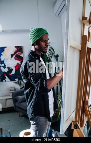 Concentré homme noir paisible peinture sur un chevalet à l'intérieur de son désordonné appartement désordonné.Un tableau abstrait sur le mur derrière. Banque D'Images