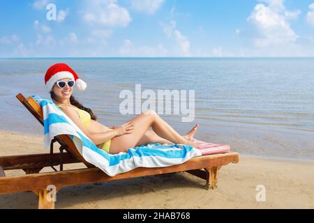 jeune femme souriante dans un chapeau de père noël et des lunettes de soleil couchant sur une chaise longue Banque D'Images