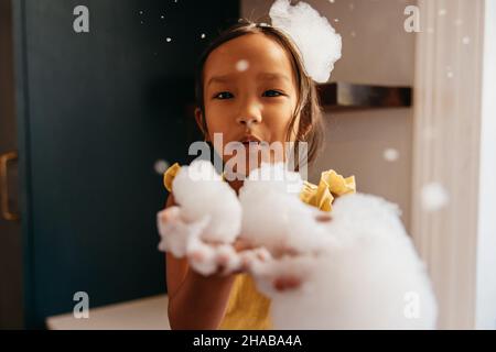 Douce petite fille soufflant des bulles de savon à la maison.Adorable jeune fille regardant l'appareil photo avec de la mousse blanche sur ses mains et sa tête. Banque D'Images