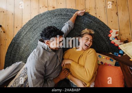 Vue en grand angle d'un couple marié riant avec joie en étant allongé dans l'aire de jeux de leurs enfants.Deux jeunes parents s'amusent les uns avec les autres à la maison. Banque D'Images