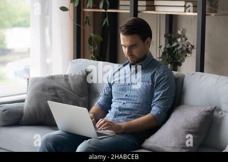 Un homme sérieux et concentré s'assoit sur un canapé à la maison avec un ordinateur portable Banque D'Images