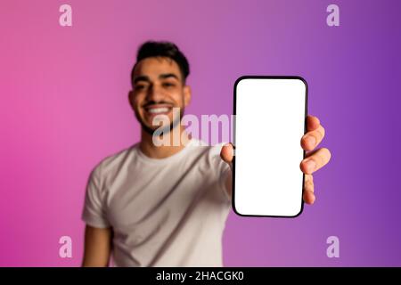 Jeune homme arabe souriant montrant un smartphone avec un écran vierge, recommandant un site Web ou une publicité dans un néon, maquette Banque D'Images