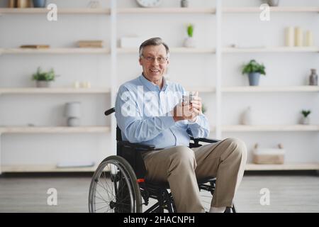 Homme positif âgé ayant une déficience assis en fauteuil roulant, buvant du café ou du thé à la maison Banque D'Images