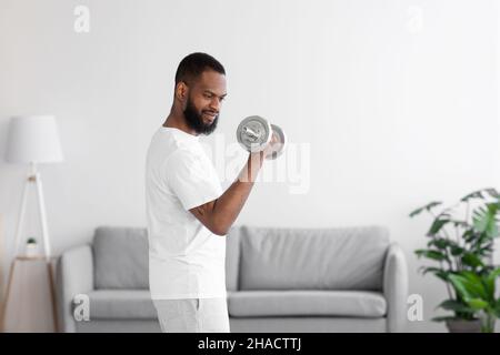 Jeune homme afro-américain barbu concentré en vêtements blancs levant des haltères Banque D'Images