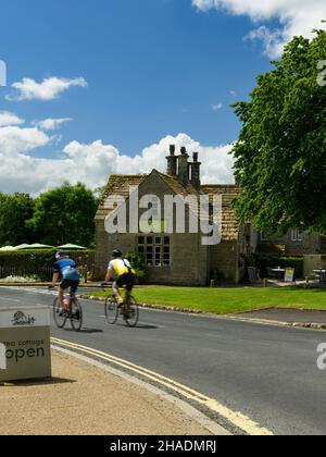 2 cyclistes sur la route de campagne passant par le beau café de salon de thé de chalet dans le pittoresque village rural ensoleillé - B6160, Bolton Abbey, Yorkshire Dales, Angleterre, Royaume-Uni. Banque D'Images