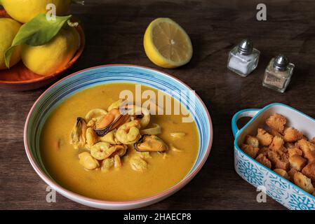 Soupe de fruits de mer à base de crevettes, moules, palourdes, merlu et calmar,servi avec du pain frit et des citrons, sur une table en bois sombre. Banque D'Images