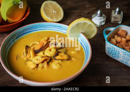 Soupe de fruits de mer à base de crevettes, moules, palourdes, merlu et calmar,servi avec du pain frit et des citrons, sur une table en bois sombre. Banque D'Images