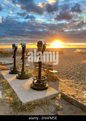Trois ensembles de jumelles à pièces debout sur une plage au coucher du soleil avec des vagues écrasant et le soleil bas dans l'arrière-plan. Banque D'Images