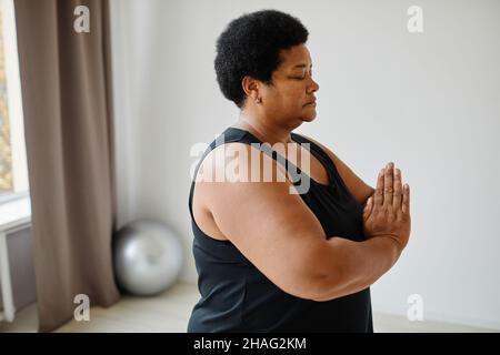 Vue latérale portrait d'une femme âgée noire méditant avec les yeux fermés tout en faisant du yoga à l'intérieur, espace de copie Banque D'Images